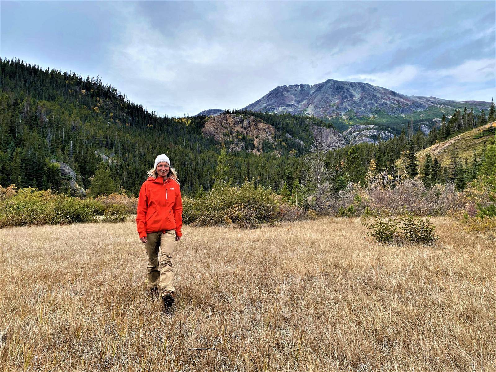 A woman hiking towards the camera on a meadow with trees and mountains in the background