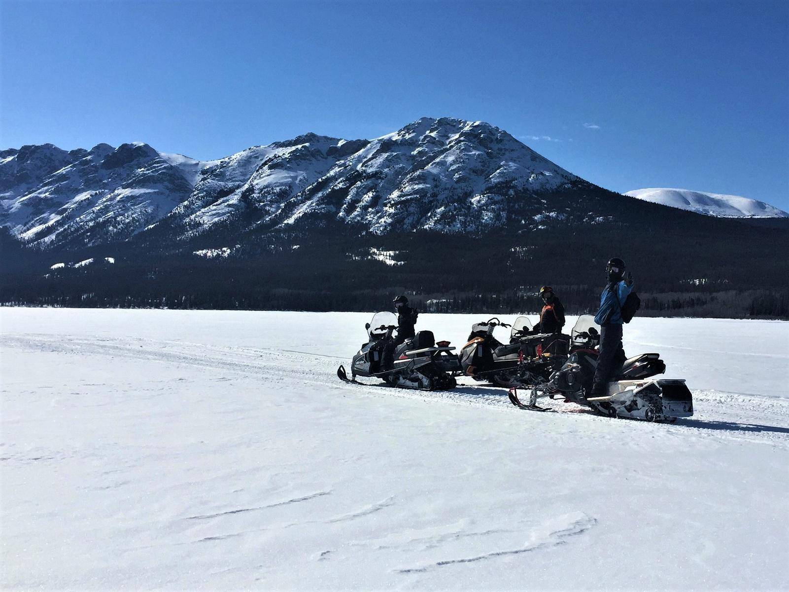 3 people riding skidoos on the frozen lake towards the snowy mountains