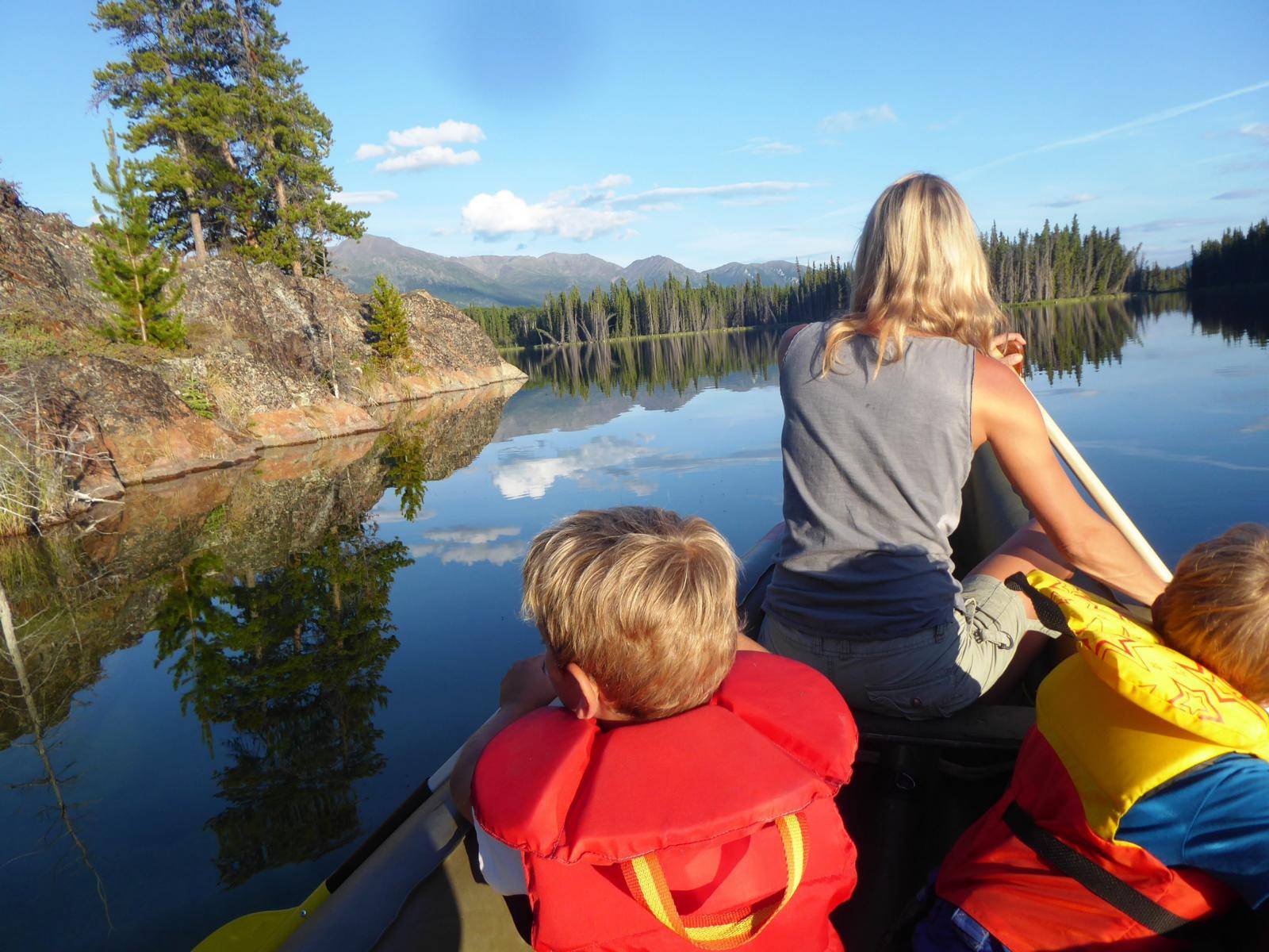 View from inside a kayak with a woman and two children paddling over a calm lake towards some trees with mountains in the background