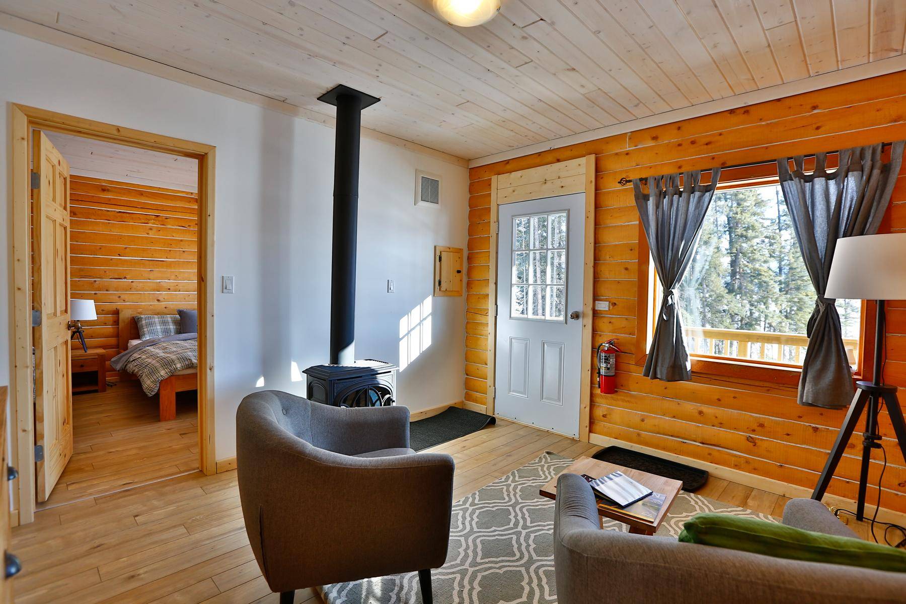 livingroom of a cabin with stove and cozy chairs, bedroom in back